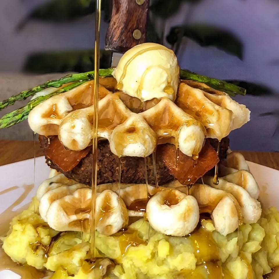 Towering burger topped with waffles, asparagus, onion rings, and ice cream, drizzled with syrup over mashed potatoes.